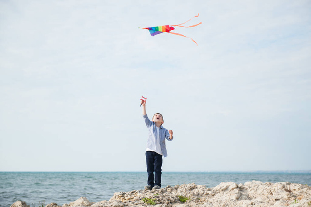 active happy little boy holding flying colorful kite flying in air standing on rock sea shore on summer sunny day - Fotoğraf, Görsel