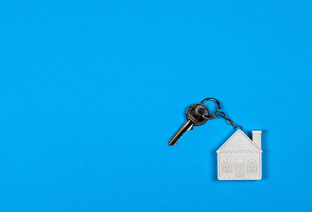 Beyaz ev şeklinde bir anahtarlık ve mavi arka planda küçük metal bir anahtar. Satın alma, satma, emlak kiralama, ipotek, komşuluk, ev - Fotoğraf, Görsel