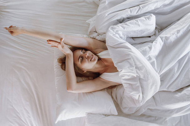 Κάτοψη της χαρούμενης ξύπνιας νεαρής ενήλικης γυναίκας, που περνά το πρωινό της στο άνετο υπνοδωμάτιο, ξαπλωμένη στο κρεβάτι, τεντωμένη και χαμογελαστή, ξεκουράζεται στο σπίτι το Σαββατοκύριακο - Φωτογραφία, εικόνα