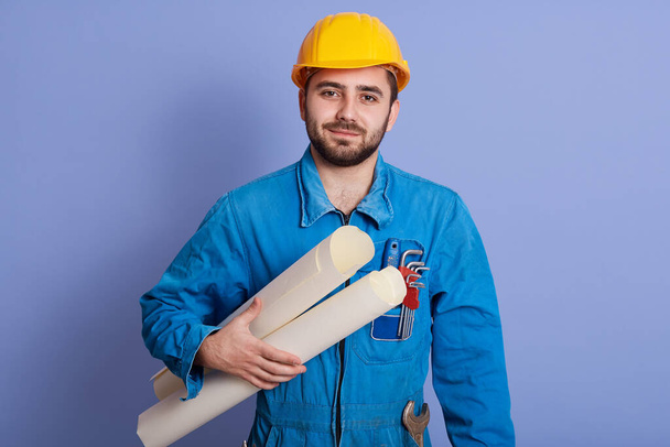 Indoor picture of calm peaceful young bearded engineer standing isolated over blue background in studio, holding papers with plan of project, having ordinary working day. People and work concept. - Φωτογραφία, εικόνα