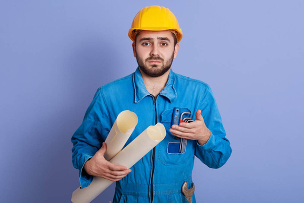 Photo of confused indignated young engineer having helpless facial expression, explaining, making gesture, wearing yellow helmet and blue uniform, being upset. People and working day concept. - Φωτογραφία, εικόνα