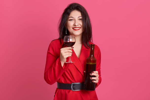 Cheerful attractive darkhaired young woman with bright makeup, holding bottle of wine and glass, lady with toothy smile celebrating something, girl posing isolated over pink studio background. - Foto, Imagen