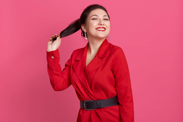 Picture of cheerful slim magnetic female smiling sincerely, having beautiful smile, holding her ponytail, wearing red jacket and black belt, being in high spirits. People and emotions concept. - Fotó, kép