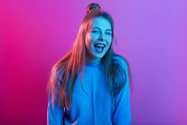 Indoor shot of magnetic charming young female opening mouth widely, having make up, laughing sincerely, having bun, standing isolated over neon lighted background. People and emotions concept. - Fotó, kép