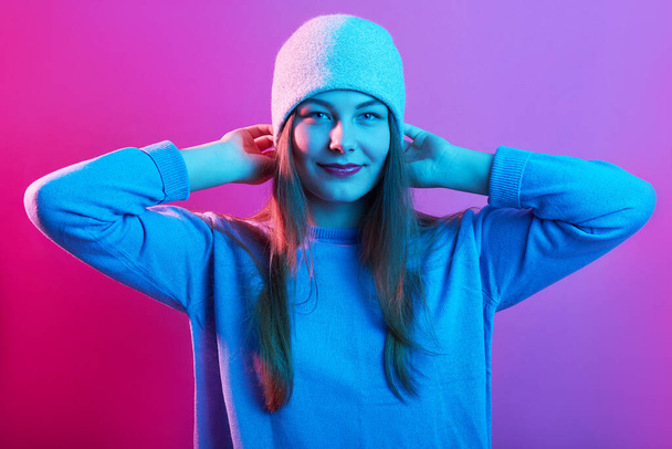 Picture of young adorable girl wearing sweater and cap, standing isolated over pink neon background, looking at camera with charming happy smile, keeping her hands behind his head, expresses happyness - Photo, image