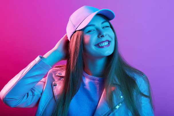 Fashionable woman stands smiling, having fun in stylish outfit. Happy beautiful girl wearing cap and leather jacket. Cheerful young model touching her head and lookins smiling directly at camera. - Photo, image