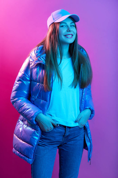 Close up portrait of beautiful woman in fashion pose looking aside in winter clothing, lady wearing jeans, cap and puffer jacket, smiling girl with hands in pocket, girl looks happy and satisfied. - Fotó, kép