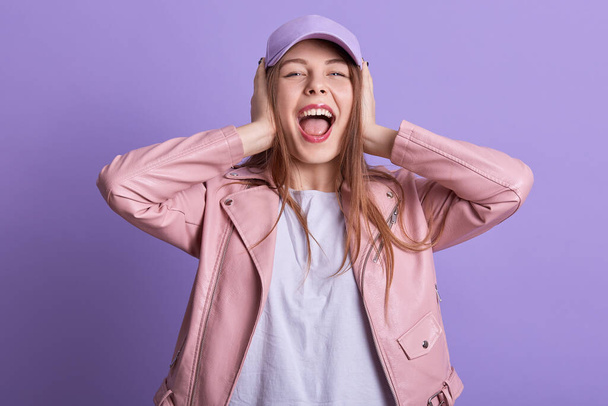 Attarctive woman in leather pink jacket covering ears and feeling irritation from loud repairing works, expressing dislike, looking directly at camera, screaming something. People emotions concept. - 写真・画像