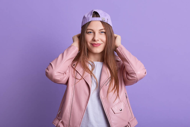 Close up portrait of pretty young woman wearing cap, white shirt and pink leather jacket, attarctive female posing isoalted over lilac studio background with hands on her head and looking at camera. - Fotoğraf, Görsel