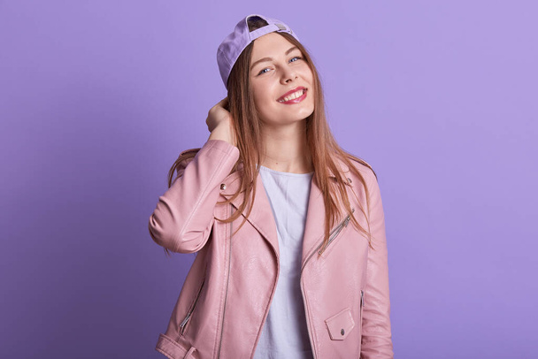 Image of pretty fair haired young female wearing pink leather jacket, white t shirt and lilac cap, having beautiful smile, looking directly at camera, standing isolated over blue background in studio. - 写真・画像