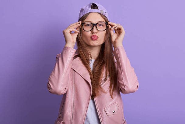Horizontal portrait of joyful adorable good looking fair haired girl protruding lips, looking directly at camera, putting on spectacles, being trendy, wearing leather jacket, cap and t shirt. - Fotografie, Obrázek