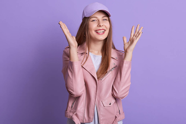 Indoor portrait of young teenager raising hands, having good news, smiling sincerely, looking directly at camera, wearing pink leather and cap, standing isolated over lilac background in studio. - Valokuva, kuva