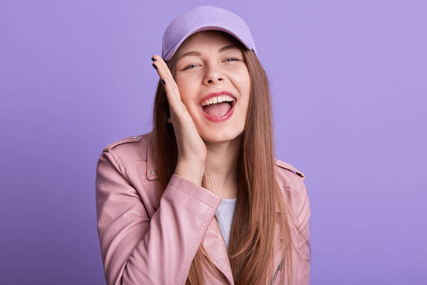 Close up portrait of cheerful sincere magnetic posing isolated over lilac background in studio, opening mouth widely, putting one hand on cheeck, laughing, having fun, feeling good. Emotions concept. - Valokuva, kuva