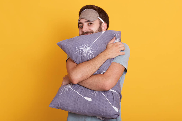 Horizontal shot of man with bare hugging pillow with satisfied facial expression isolated over yellow background, male with sleeping mask on forehead, wants to sleep, looking at camera, wears t shirt. - 写真・画像