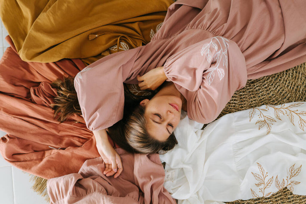 Sensual young woman in pink dress lying on the floor over composition of differently colored dresses of the same fashion. Posing for a photo. - Photo, image