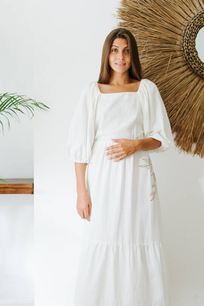 Pretty happy young woman with inocent look in a light white summer dress posing for a photo in a tropical style room. Her left hand on the belly. - Фото, изображение