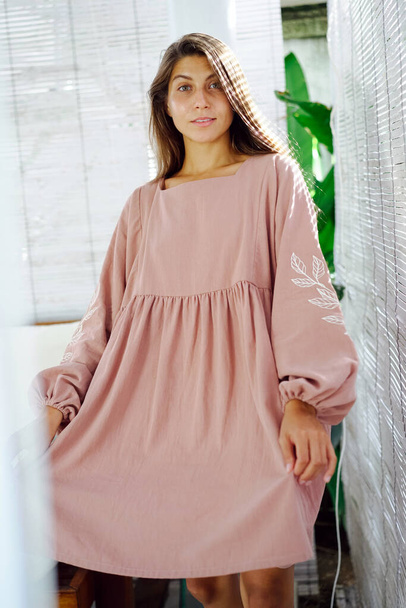 Happy young woman in pink midi dress standing in a bright room, posing, looking at camera. Pulling, holding dress with one hand. - Fotoğraf, Görsel