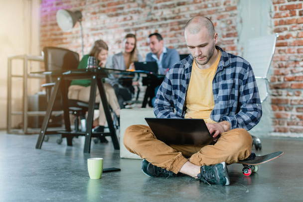 Hip worker in a startup coding with laptop sitting on skateboard - Фото, изображение