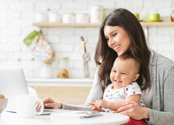 Szczęśliwa kobieta pracuje na laptopie z dzieckiem w kuchni - Zdjęcie, obraz