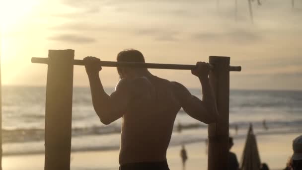 Młody muskularny mężczyzna podciąga się na poziomym barze na plaży na tle zachodu słońca - Materiał filmowy, wideo