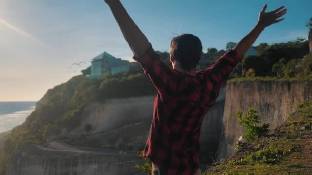 Young man traveler raising his hands high on top of the mountain above beautiful landscape on golden sunset - Video