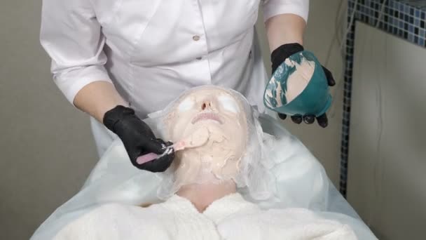 Foto ravvicinata di una giovane donna che riceve un trattamento facciale nel salone di bellezza. Una cliente donna in una clinica di bellezza. estetista in guanti neri applicando con spatola maschera di argilla sul viso coperto di garza. 4K video
 - Filmati, video