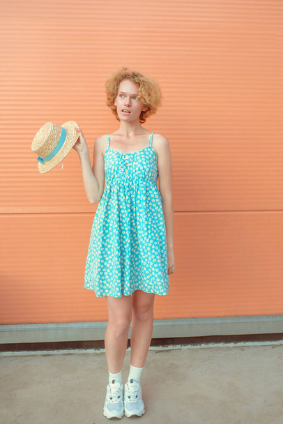 młoda, wesoła, kręcona ruda kobieta w niebieskiej sukience, trzymająca słomiany kapelusz w dłoni na beżowym tle. Zabawa, lato, moda, koncepcja młodzieży - Zdjęcie, obraz