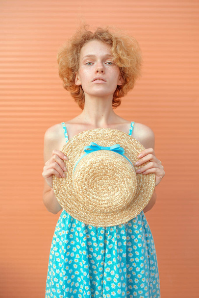 young cheerful curly redhead woman in blue sundress holding straw hat in her hand on beige background. Fun, summer, fashion, youth concept - Fotó, kép