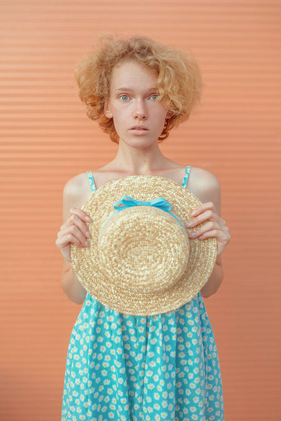 young cheerful curly redhead woman in blue sundress holding straw hat in her hand on beige background. Fun, summer, fashion, youth concept - Fotografie, Obrázek
