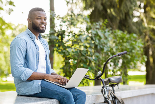 ノートパソコンを持って公園に座っているアフリカ系の男、屋外でエッセイを書く - 写真・画像