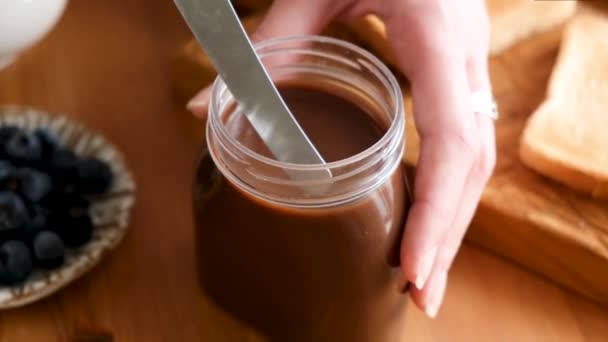Chocolate nut spread or butter in jar. Person taking dollop of chocolate nut butter with table knife to smear it on bread toast - Filmagem, Vídeo