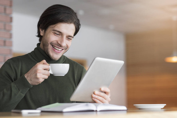 Happy young man reading emails while drinking coffe at cafe - Фото, изображение