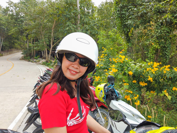 Jovencita tailandesa con casco. Detrás de ella, una motocicleta verde y un fondo tropical
 - Foto, Imagen