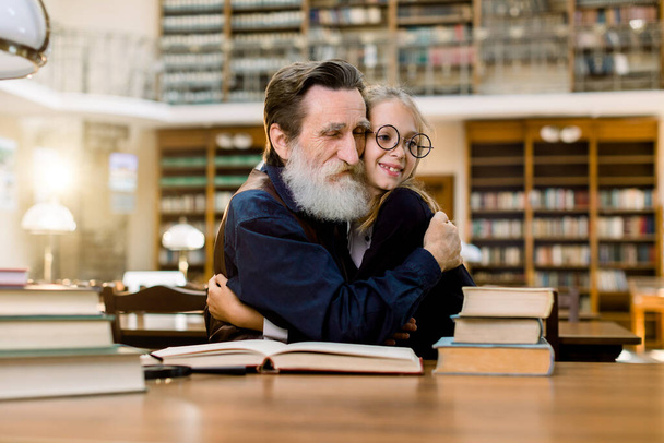 Grandfather and granddaughter or senoir teacher and pupil student, sitting at the table and hugging each other, in old vintage city library. Reading, education concept. Happy international book day - Foto, Imagem