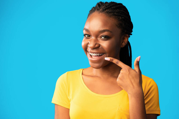 Africano senhora no aparelho apontando o dedo para os dentes, fundo azul
 - Foto, Imagem