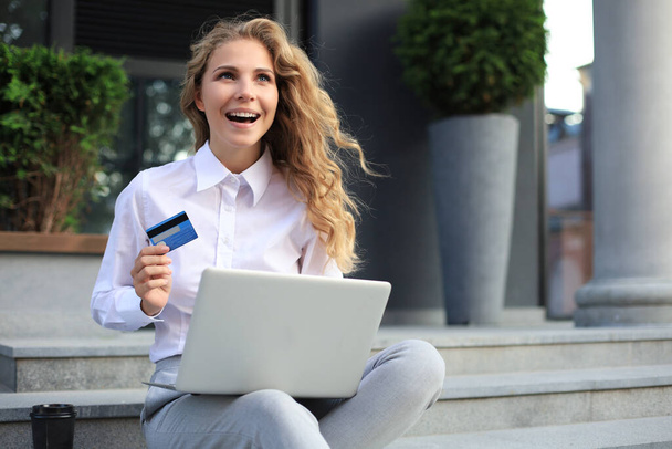 Online shopping concept. Young blonde woman holding a credit card and doing online payment with laptop outdoors - 写真・画像