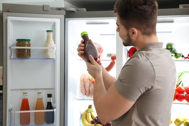 Young man with bottle of sauce near open refrigerator indoors - Фото, изображение