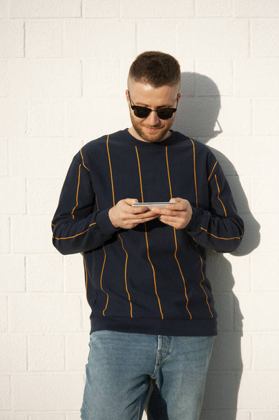Guy in casual wear with mobile phone. Young caucasian man standing at outdoor white wall and see videos or playing video games with smartphone. Leisure activity with technology devices. - Foto, Bild