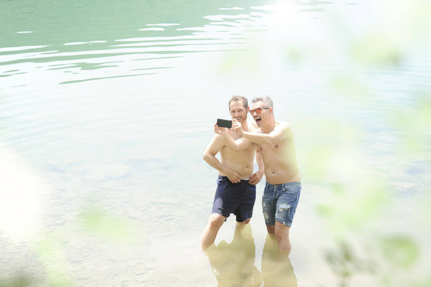 Dos hombres parados juntos al aire libre tomando una selfie y sonriendo. Los mejores amigos tomando una selfie mientras están de pie en el agua del lago en un día de verano. Hombres con pantalones vaqueros cortos. Pecho desnudo para ambos
. - Foto, Imagen