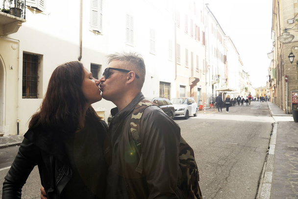 Couple of travelers are kissing in the street. Man and woman are wearing in casual clothing. Leather jacket for both. Sunglasses for him. Lifestyle theme. - Foto, imagen