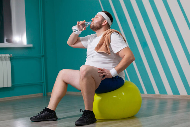 Overweight man is sitting on a fitness ball exhausted after a hard training in group fitness classes. Fat man is drinking water with a towel on his shoulder - Photo, image