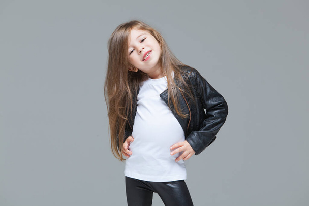 Baby girl with long hair in black leather jacket and leggings is standing in the studio on grey background - 写真・画像