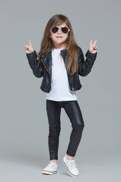 Baby girl with long hair in black leather jacket and leggings is posing in the studio in modern sunglasses on grey background - Foto, immagini
