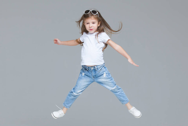 Kid girl preschooler in blue jeans, white t-shirt and sunglasses is jumping with raised hands isolated on grey background - Photo, Image