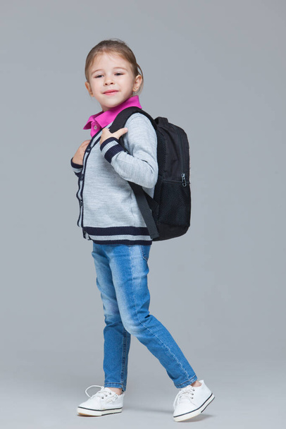 Primary school girl in jeans and uniform jacket is wearing backpack. Isolated on grey background - Photo, image