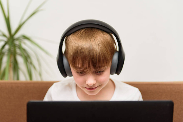 Εφηβικό αγόρι κάθεται στο φορητό υπολογιστή με ακουστικά στο φως φόντο. Σύγχρονη εκπαιδευτική έννοια. - Φωτογραφία, εικόνα