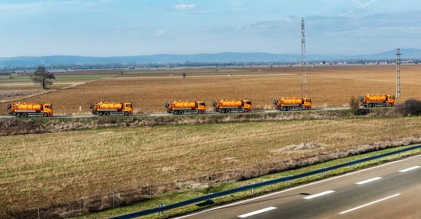 Comboio ou Caravana de caminhões-tanque de laranja em uma rodovia sinuosa através da paisagem rural
 - Foto, Imagem