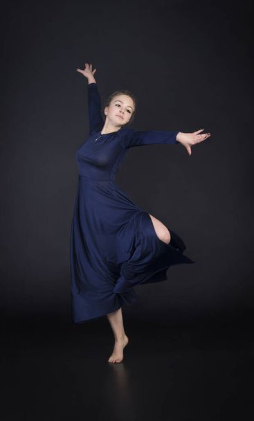 A girl in a blue dress with light hair is dancing a modern ballet. Studio shooting on a dark background. - Фото, зображення