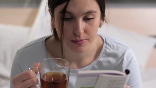 Dziewczyna czyta książkę. Młoda atrakcyjna biała kobieta siedzi w łóżku wieczorem i czyta książki i pije czarną herbatę przed snem. Zbliżenie - Materiał filmowy, wideo
