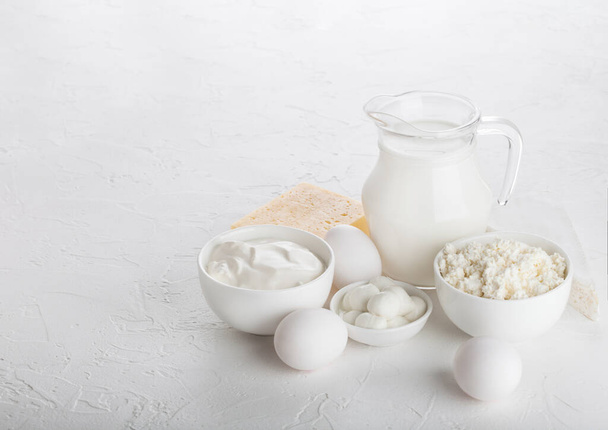 Φρέσκα γαλακτοκομικά προϊόντα σε λευκό φόντο πίνακα. Γυάλινο βάζο με γάλα, μπολ με ξινή κρέμα και τυρί cottage και μοτσαρέλα. Αυγά μέσα και τυρί.  - Φωτογραφία, εικόνα